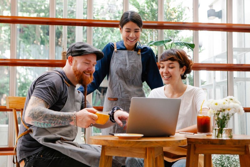 Three café staff in aprons smiling at a laptop on a café table.